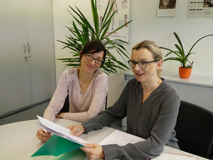 Zwei Gutachterinnen des Medizinischen Dienstes Mecklenburg-Vorpommern beraten über ein fachliches Thema. Sie schauen kurz auf, um in die Kamera zu lächeln.
